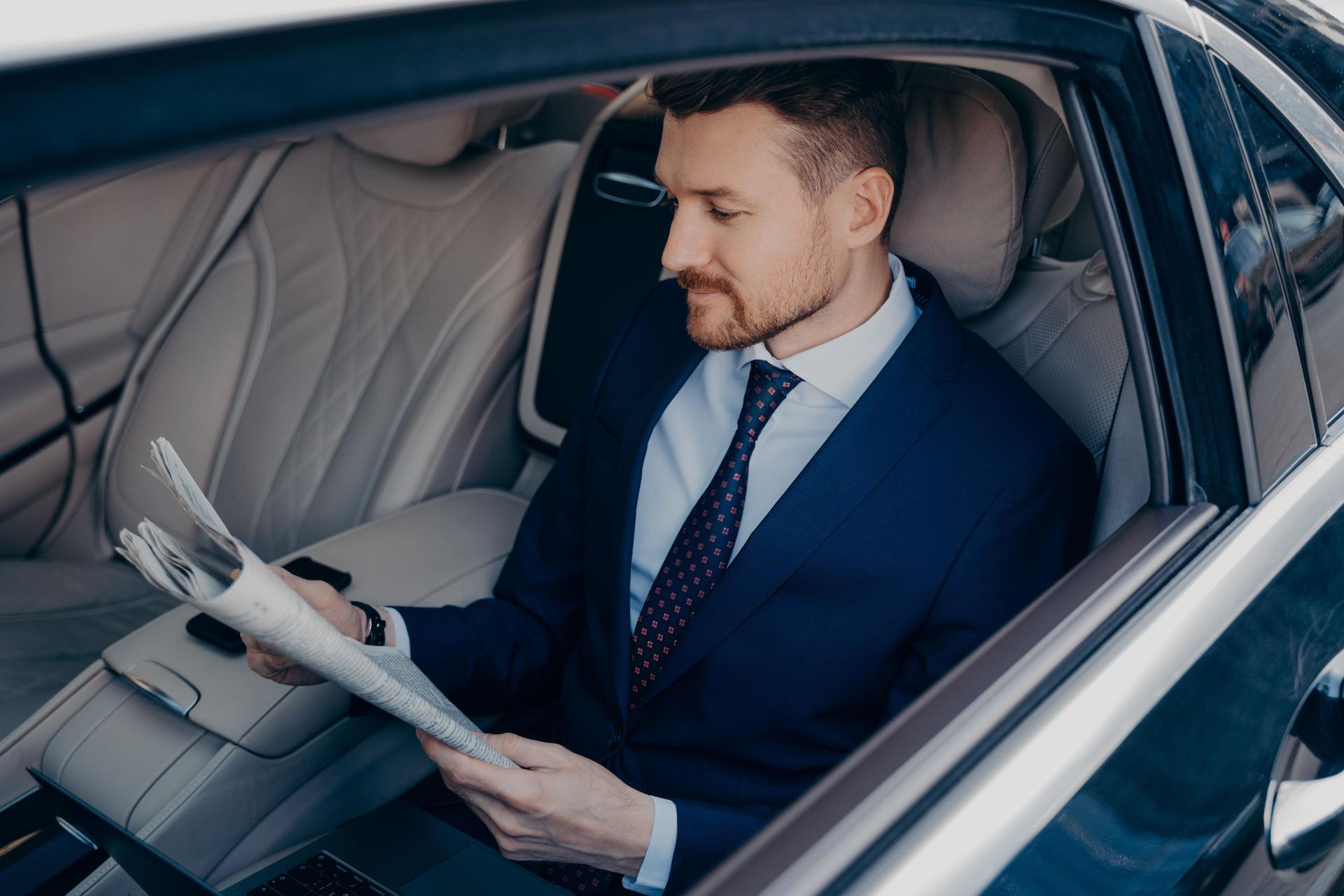 Thoughtful executive manager in formal suit reading newspaper in backseat of limousine